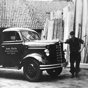 Foto em preto e branco do primeiro empregado da Dolle em pé ao lado de um caminhão vintage carregado de madeira.