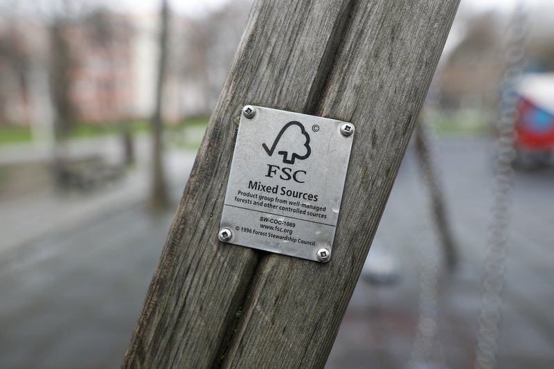 Um zoom de uma estrutura de madeira com uma placa metálica FSC (Forest Stewardship Council) fixada, indicando o uso de materiais provenientes de fontes responsáveis, com um parque desfocado ao fundo.
